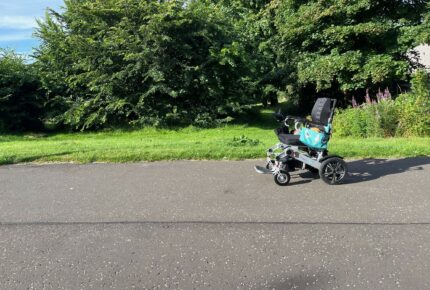 Powerchair from the side on a tarmac path with grass and trees in the background.